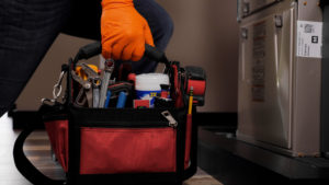 Gloved hand holding a toolbag in front of furnace unit